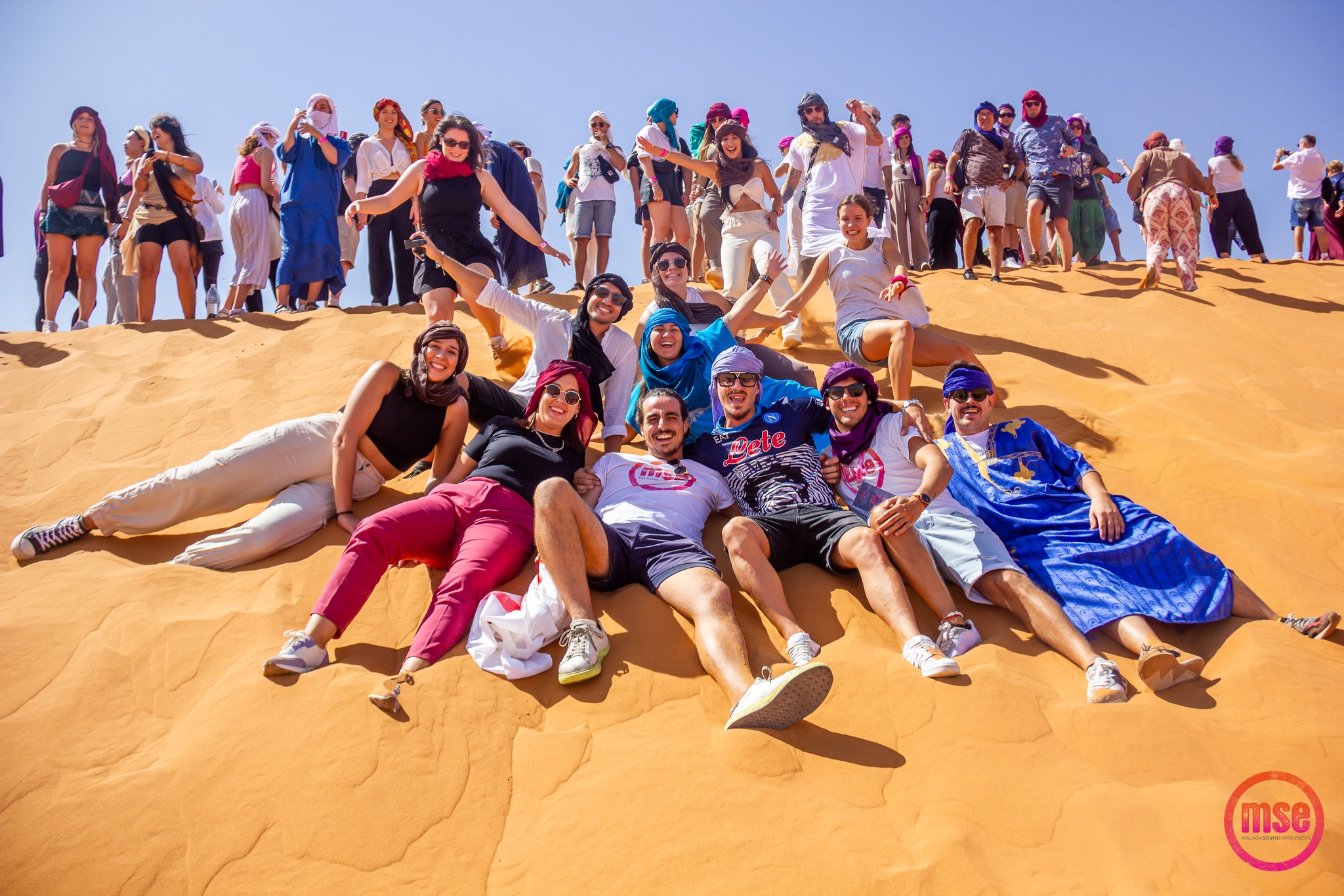 Grupo de turistas disfrutando en las dunas de arena del Sahara durante un viaje organizado por Malaga South Experiences. Los participantes están sentados y deslizándose por la pendiente de una gran duna, luciendo sonrisas y expresiones de alegría. Visten ropa casual y colorida, algunos con pañuelos tradicionales en la cabeza, bajo un cielo azul claro y sin nubes. La vitalidad y el ambiente festivo se reflejan en sus posturas relajadas y brazos extendidos.